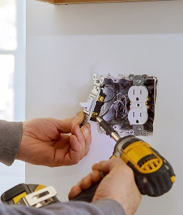 Main d'un électricien installant un interrupteur et une prise électrique murale avec un tournevis et une perceuse.