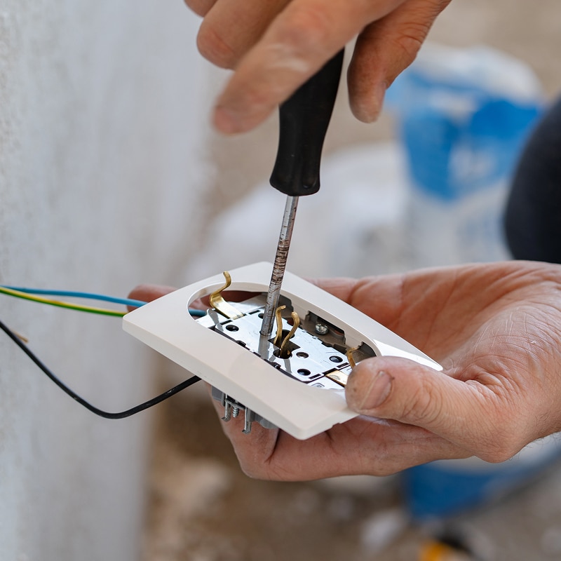 Gros plan sur un électricien vissant une prise électrique murale avec un tournevis. Fils bleus, jaunes et noirs connectés.